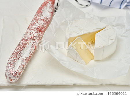 A round head of soft Camembert cheese with white mold, one triangular slice cut off, the cheese lies on crumpled white wrapping paper A round head of soft Camembert cheese with white mold, one triangular slice cut off, the cheese lies on crumpled white wrapping paper 136364011