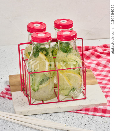 Glass bottles with red lids, filled with detox water containing cucumber and lemon slices and mint leaves, white table 136364052
