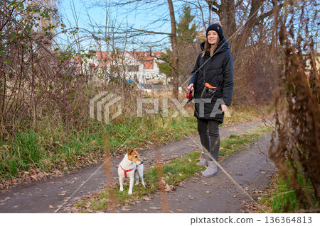 Woman walking small dog on leash along path surrounded by trees Woman walking small dog on leash along path surrounded by trees 136364813