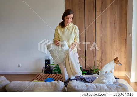 Woman standing in living room holding pillow and blanket while tidying sofa 136364815