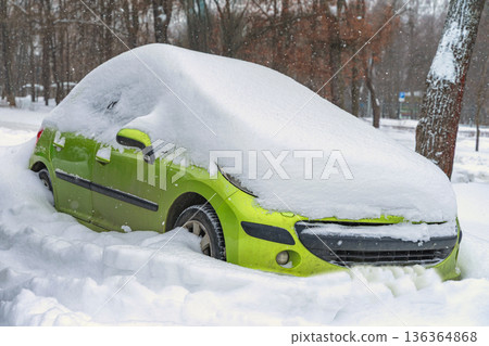 Green car parked on city street buried in snow after blizzard, windshield hidden by snow, parking blocked. Concept of snow disruption, winter transport, road safety, insurance claim 136364868