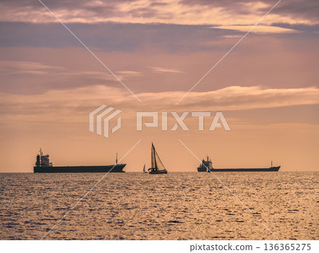 Sunset Seascape With Cargo Ships And A Sailboat On Calm Ocean Horizon During Golden Evening Sunset Seascape With Cargo Ships And A Sailboat On Calm Ocean Horizon During Golden Evening 136365275