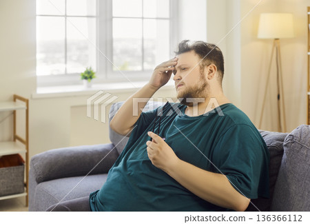 Stressed overweight man rubbing forehead on sofa at home, holding eyeglasses 136366112