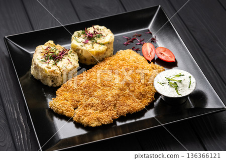 Crispy breaded schnitzel with mashed potatoes and creamy sauce on black plate, dark table 136366121