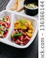 Takeout box with chicken broth soup, toast, fresh salad, potato wedges and sausage 136366144