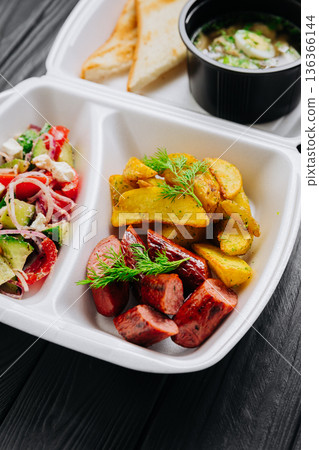 Takeout box with chicken broth soup, toast, fresh salad, potato wedges and sausage 136366144