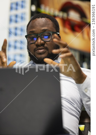 Black man participating in online video call while sitting on bench, confident street style and relaxed posture fitting naturally into the modern downtown area with vibrant active city life. 136366191