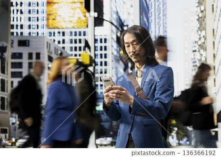 Portrait of asian businessman scrolling on phone among pedestrians in a downtown street, showing multicultural crowd, blurred motion and realistic routine of active urban environment. 136366192