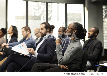 Black young female listening to presentation speech with diverse people in audience, business conference event with speaker. Chairs fill auditorium as participants attend professional summit. 136366195