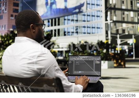 Young black adult programming binary code on terminal window, sitting on a bench downtown and working on IT with his laptop. Professional attitude in a dynamic city life avenue. Young black adult programming binary code on terminal window, sitting on a bench downtown and working on IT with his laptop. Professional attitude in a dynamic city life avenue. 136366196