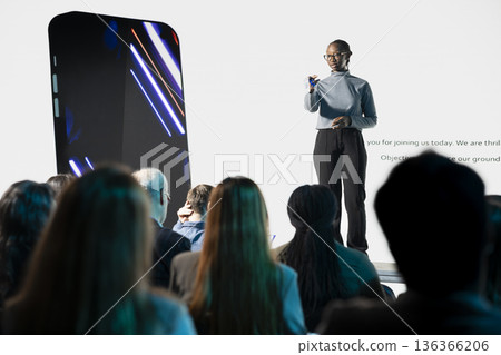 African american girl with microphone introducing new smartphone on stage, release announcement with promotion visuals. Advertising new launch for investment deal during convention event. African american girl with microphone introducing new smartphone on stage, release announcement with promotion visuals. Advertising new launch for investment deal during convention event. 136366206