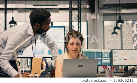 Diverse work team in a multinational company office discussing forecasting and resource allocation. Visual data and reports displayed on laptop support strategic planning and goals achievement. 136366211