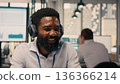 Cheerful black man listening to audio playlist on wireless headphones, reviewing charts and graphs on laptop. Relaxed worker multitasking with fun music, stress relief in modern office. 136366214