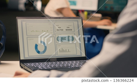 Male office worker solving tasks independently on laptop at desk, reviewing visual data, charts and graphs. Studying forecasting reports for productivity and global business planning. 136366224