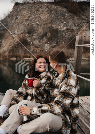 Couple enjoys warm drinks while seated on a dock next to a lake during a cold day together 136366233