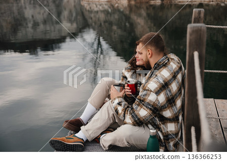 Couple enjoys warm drinks together by the water on a cold day near a quiet shoreline 136366253