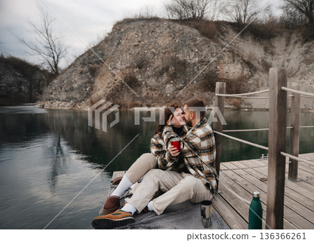 Couple enjoys time together by the water on Valentines Day while sharing warm drinks and smiles, wrapped in cozy sweaters 136366261