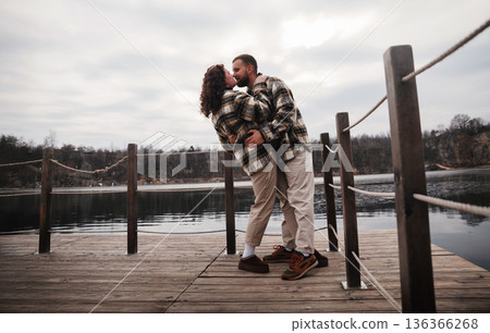 Couple enjoying a moment of love while standing on a wooden pier by the water on a cloudy day 136366268
