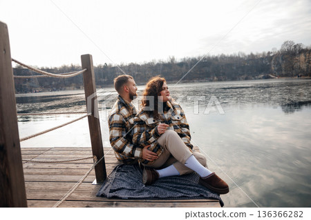 Couple enjoys time by the lake sitting close while wrapped in blankets on a chilly day 136366282