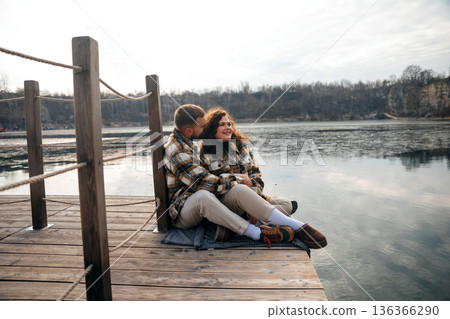 Couple sits on dock enjoying time together on a chilly day by the water for Valentines Day celebration 136366290