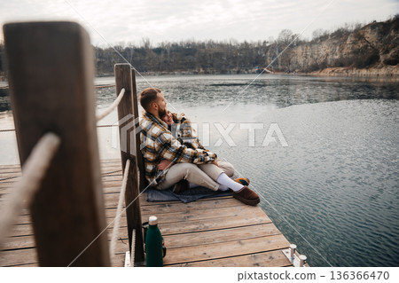 Couple sits on wooden dock by the water enjoying each other company on a chilly day during Valentin season 136366470