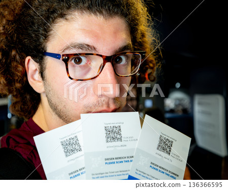 Berlin, germany, august 12, 2023. Young man with glasses holding multiple qr-code tickets up to camera, surprised expression while checking digital event passes indoors Berlin, germany, august 12, 2023. Young man with glasses holding multiple qr-code tickets up to camera, surprised expression while checking digital event passes indoors 136366595