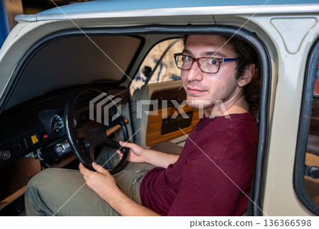 Berlin, germany, august 12, 2023. Young man in glasses behind the wheel of a vintage car, smiling and looking at the camera while driving on a sunny road Berlin, germany, august 12, 2023. Young man in glasses behind the wheel of a vintage car, smiling and looking at the camera while driving on a sunny road 136366598