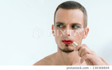 portrait of adult gay man with makeup and cosmetics on face. A handsome transgender man with bright eyeshadows paints lips with red lipstick portrait of adult gay man with makeup and cosmetics on face. A handsome transgender man with bright eyeshadows paints lips with red lipstick 136366604