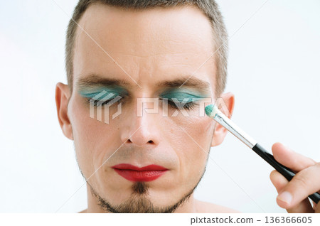 portrait of adult gay man with makeup and cosmetics on face. A handsome male with red lipstick applied eye shadow sparkle on his eyes with brush portrait of adult gay man with makeup and cosmetics on face. A handsome male with red lipstick applied eye shadow sparkle on his eyes with brush 136366605