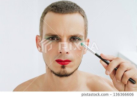 portrait of adult gay man with makeup and cosmetics on face. A handsome transgender male with red lipstick applied eye shadow sparkle on eyes with a brush on a white background portrait of adult gay man with makeup and cosmetics on face. A handsome transgender male with red lipstick applied eye shadow sparkle on eyes with a brush on a white background 136366606