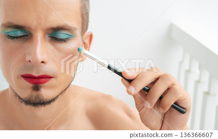 portrait of feminine gay man with makeup and cosmetics on face. A handsome young transgender male with red lipstick applied eye shadow sparkle on his eyes with a brush on a white background portrait of feminine gay man with makeup and cosmetics on face. A handsome young transgender male with red lipstick applied eye shadow sparkle on his eyes with a brush on a white background 136366607