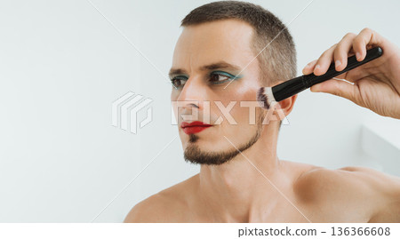 portrait of a feminine gay man with makeup and cosmetics on face. A handsome young transgender man with red lipstick on lips and painted eyes applies blush with brush and does facial contouring portrait of a feminine gay man with makeup and cosmetics on face. A handsome young transgender man with red lipstick on lips and painted eyes applies blush with brush and does facial contouring 136366608