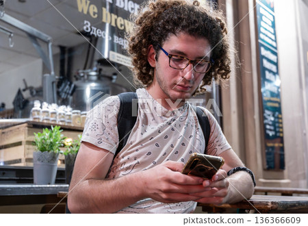 Berlin, germany, august 12, 2023. Young curly haired man with glasses texting on smartphone in urban cafe, casual gen z lifestyle, connected and focused on device Berlin, germany, august 12, 2023. Young curly haired man with glasses texting on smartphone in urban cafe, casual gen z lifestyle, connected and focused on device 136366609