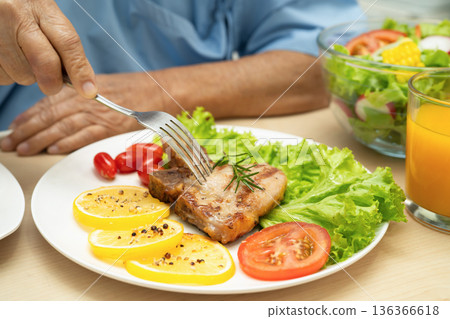 Asian senior woman patient eating pork chop stake and vegetable salad for healthy food in hospital. 136366618
