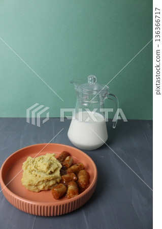 Homemade lunch, mashed potatoes, sausages in tomato red sauce, a jug of milk on a gray background. The concept of simple homemade food. Vertical Photo Side View. Homemade lunch, mashed potatoes, sausages in tomato red sauce, a jug of milk on a gray background. The concept of simple homemade food. Vertical Photo Side View. 136366717