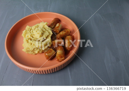 Mashed potatoes with livka with sausages in tomato sauce in a peach orange plate on a gray background with space for copyspace text. Mashed potatoes with livka with sausages in tomato sauce in a peach orange plate on a gray background with space for copyspace text. 136366719