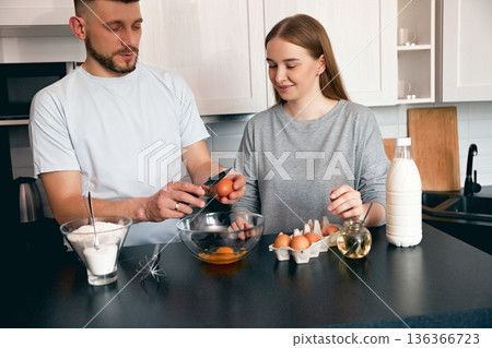 Couple prepares breakfast in a kitchen with eggs, milk, and flour on a counter while smiling and working together Couple prepares breakfast in a kitchen with eggs, milk, and flour on a counter while smiling and working together 136366723