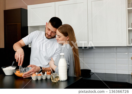 Couple cooking together in a kitchen at home while preparing a meal with eggs, milk, and other ingredients in the morning Couple cooking together in a kitchen at home while preparing a meal with eggs, milk, and other ingredients in the morning 136366726