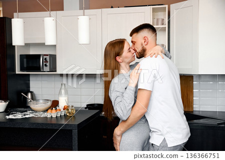Couple shares a kiss in the kitchen while preparing breakfast together during a cozy morning at home 136366751