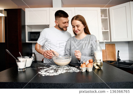 Couple cooking together in a kitchen while preparing a dish using flour, eggs, and mixing ingredients in a bowl Couple cooking together in a kitchen while preparing a dish using flour, eggs, and mixing ingredients in a bowl 136366765