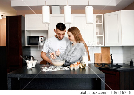 Couple makes breakfast together in modern kitchen with eggs and flour on counter while smiling at each other and mixing batter in bowl Couple makes breakfast together in modern kitchen with eggs and flour on counter while smiling at each other and mixing batter in bowl 136366766