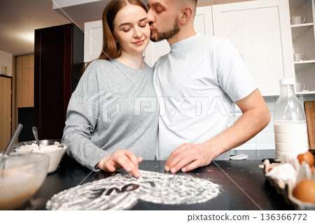 Couple prepares dessert together in kitchen while sharing a kiss and smiling in cozy indoor setting during daytime hours 136366772