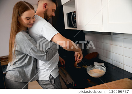 Couple cooks together in kitchen while sharing a warm moment in their home during morning hours on a weekend Couple cooks together in kitchen while sharing a warm moment in their home during morning hours on a weekend 136366787