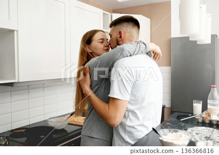 Couple embraces in kitchen while cooking together and sharing a moment in the afternoon light 136366816