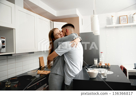 Couple shares a hug in the kitchen during a home cooking session in the afternoon, surrounded by ingredients and kitchen items 136366817