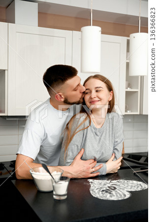 Couple shares a moment in the kitchen as one partner kisses the other while they stand near a countertop filled with dessert ingredients Couple shares a moment in the kitchen as one partner kisses the other while they stand near a countertop filled with dessert ingredients 136366823