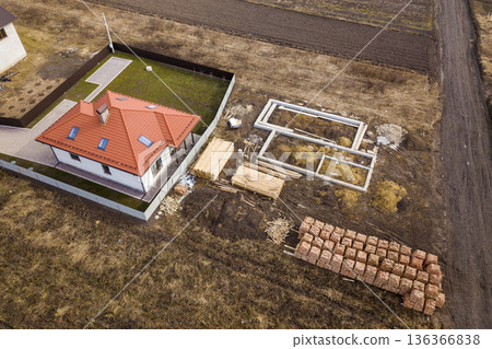Aerial view of new house roof with attic windows and building site, foundation of future house, stacks of bricks and building timber logs for construction. Aerial view of new house roof with attic windows and building site, foundation of future house, stacks of bricks and building timber logs for construction. 136366838
