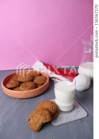 Cookies with milk for Santa Claus. Oatmeal cookies on a plate, a glass of milk, a Santa Claus hat on a gray pink background. Treat for Santa. 136367370