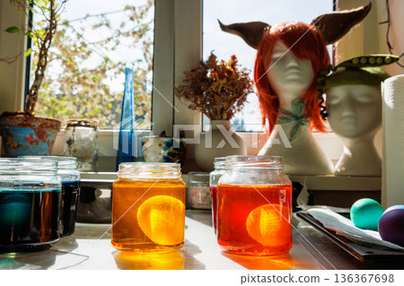 Sunlit kitchen craft scene with easter eggs soaking in a glass jars of yellow and orange food dye near a window 136367698