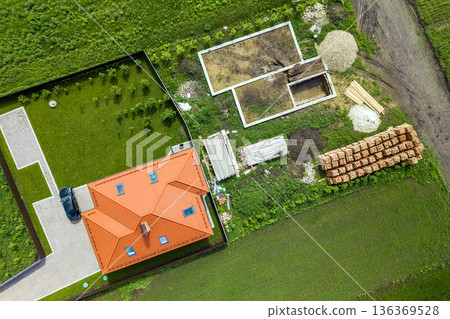 Aerial top view of a private house with attic windows on roof, paved yard with green grass lawn and building site with concrete foundation floor and stacks of yellow bricks for construction.. 136369528